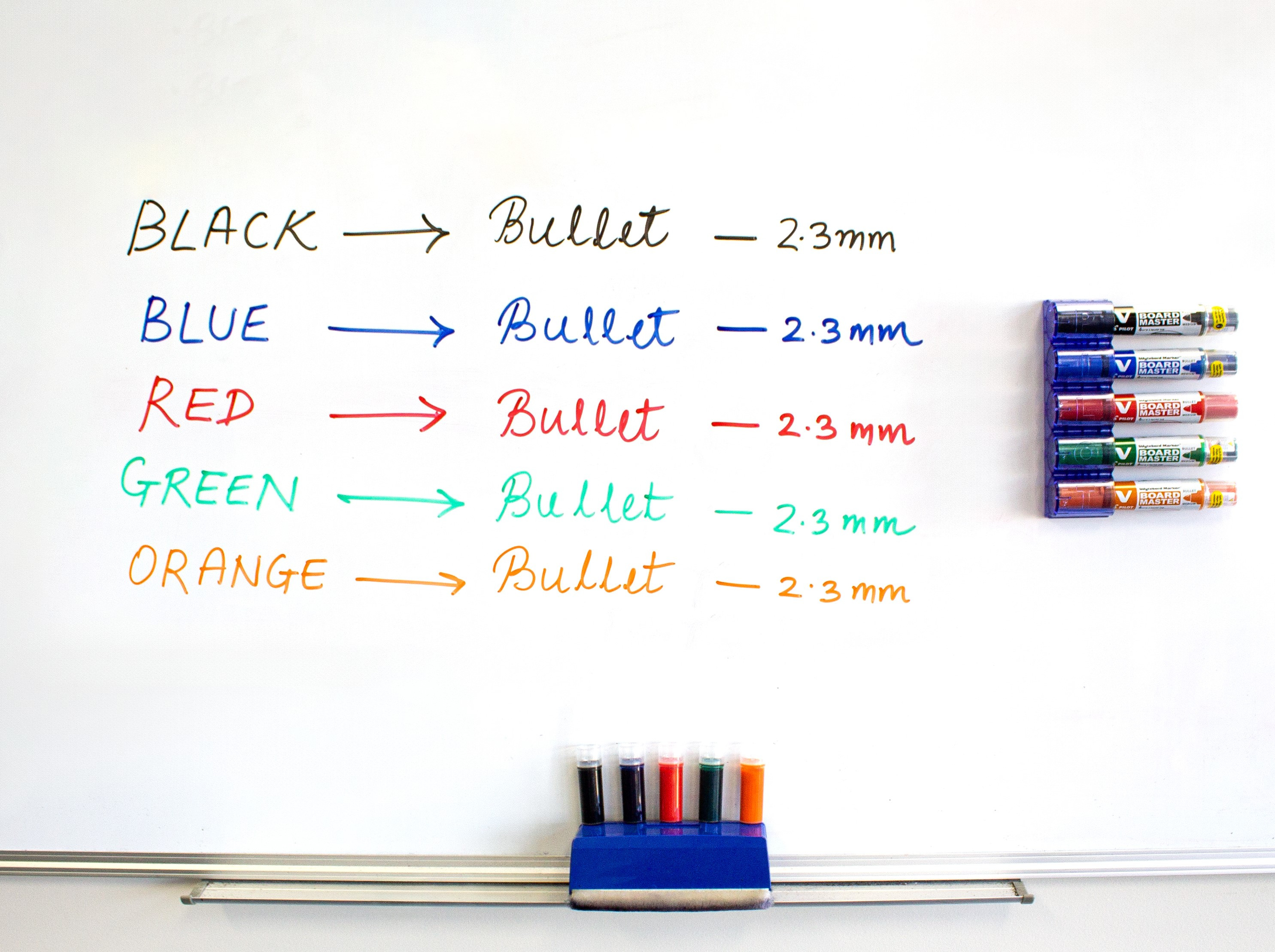 Whiteboard displaying colour-coded handwritten notes using PILOT V Board Master markers in black, blue, red, green, and orange, with corresponding markers mounted on the side and refill cartridges neatly arranged at the bottom.