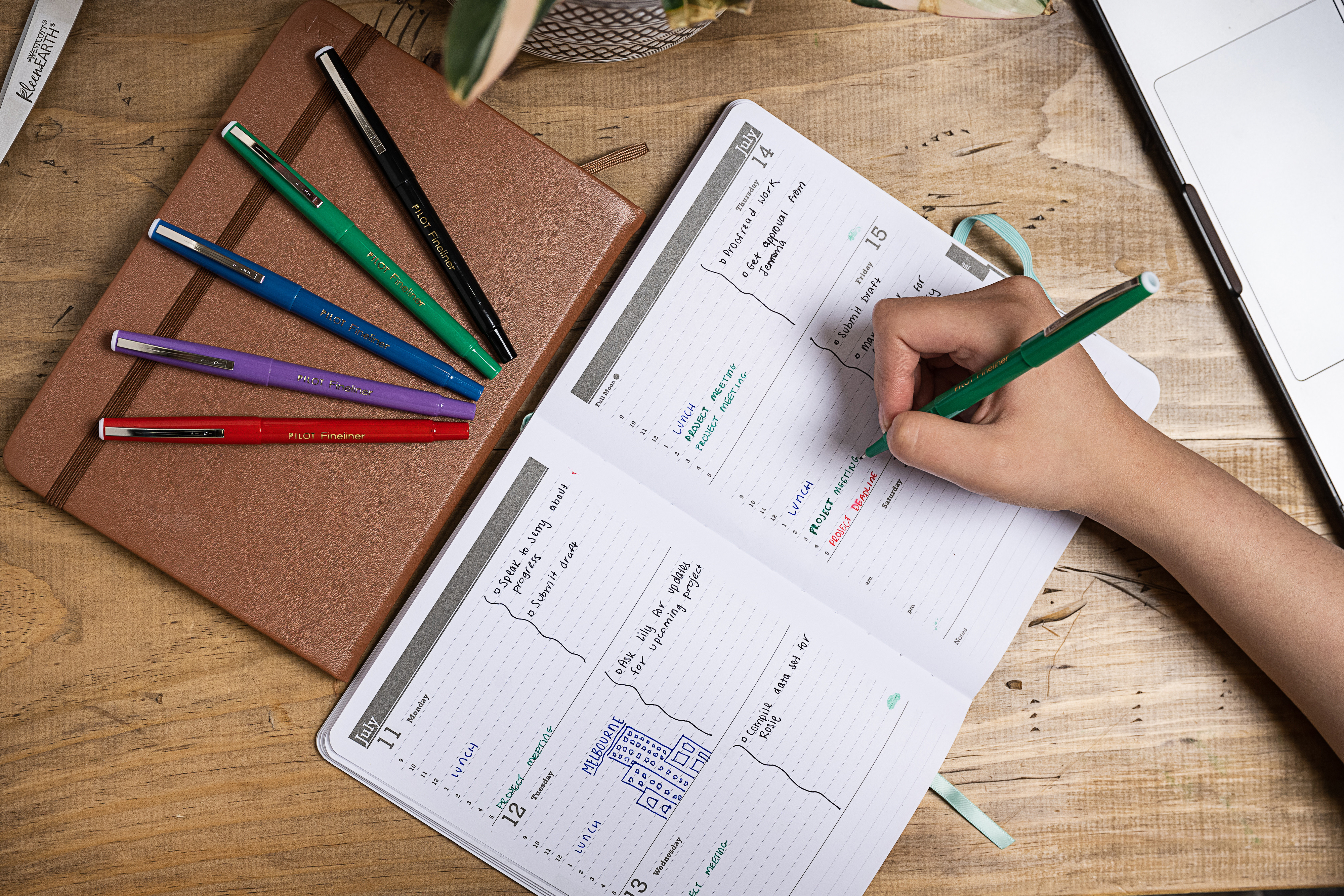 Using a green PILOT Fineliner marker to write in a bullet journal weekly spread, with colourful fineliners arranged beside a notebook on a wooden desk