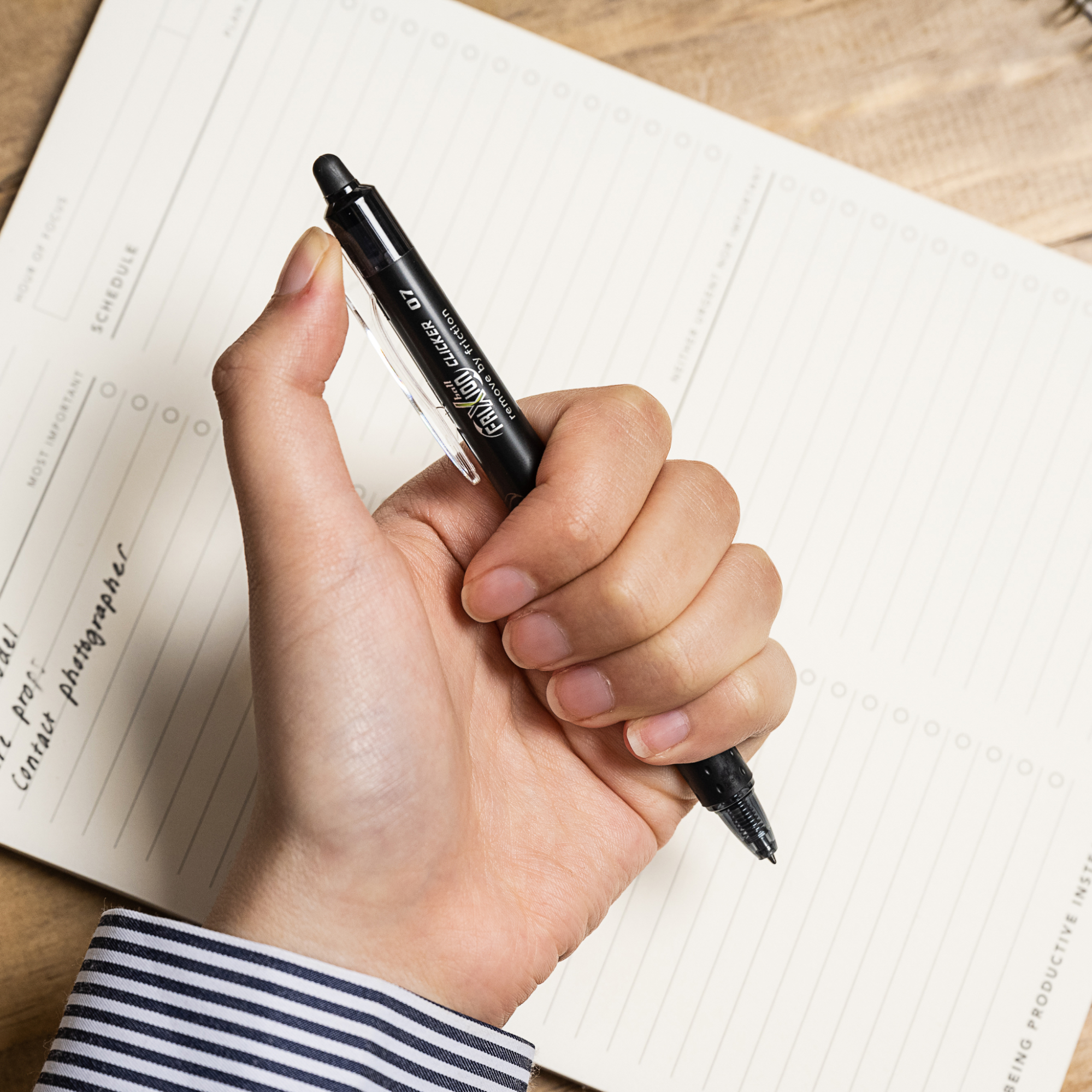 Close-up of a hand holding a black FriXion Clicker pen above a blank daily planner, a good quality pen for smooth, organised writing.