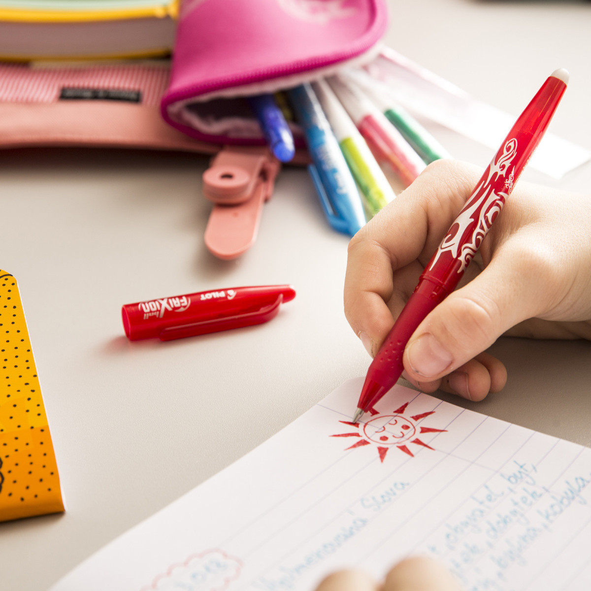 Child drawing a sun with PILOT&rsquo;s FriXion Stick Erasable Gel Pen, surrounded by a colourful array of pens, ideal for kids' creative projects