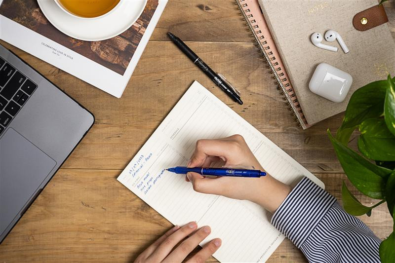 A person using a blue and black erasable FriXion pen to write their daily to do list next to their laptop
