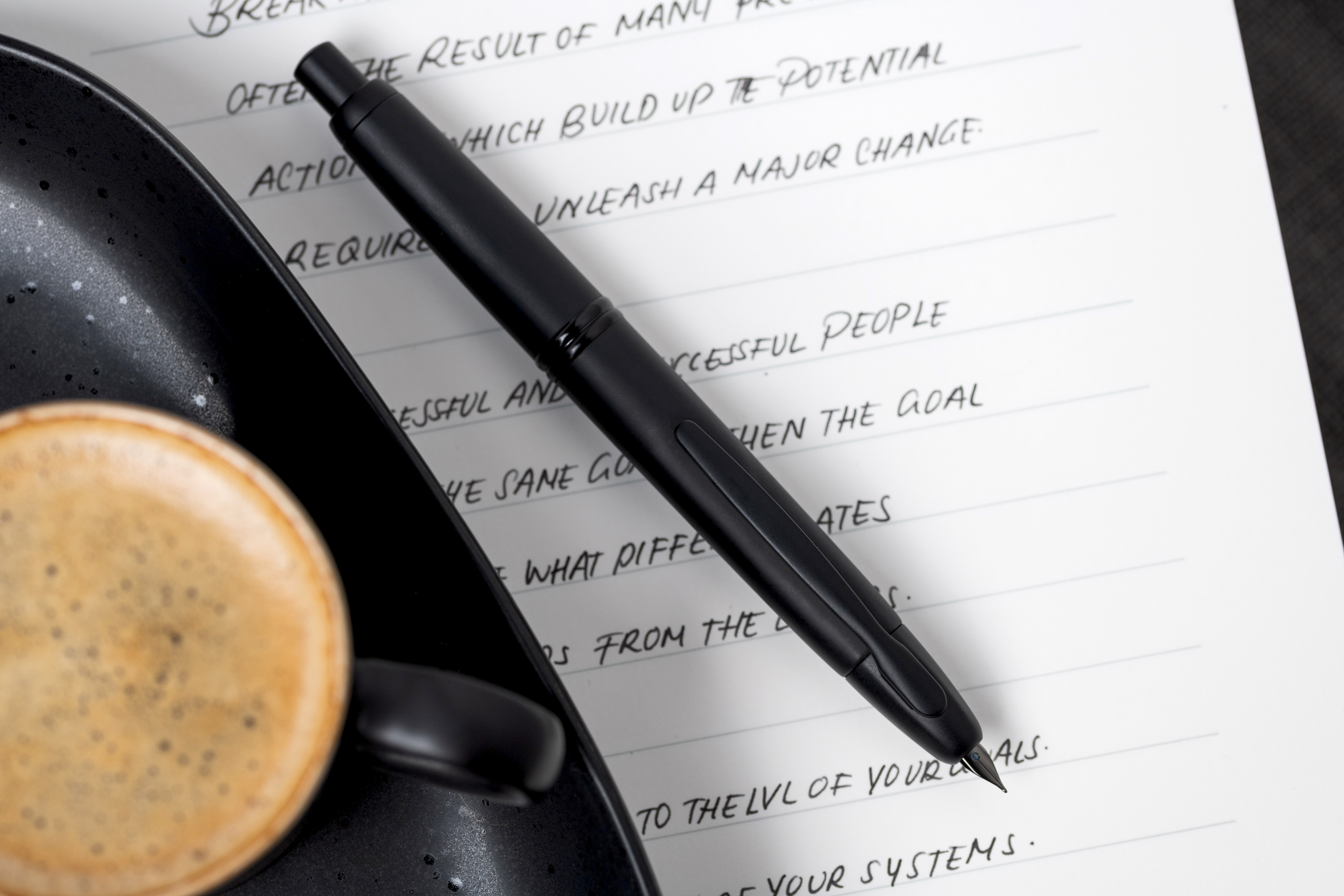 Pilot Capless Matte Black fountain pen resting on a handwritten notebook beside a cup of coffee