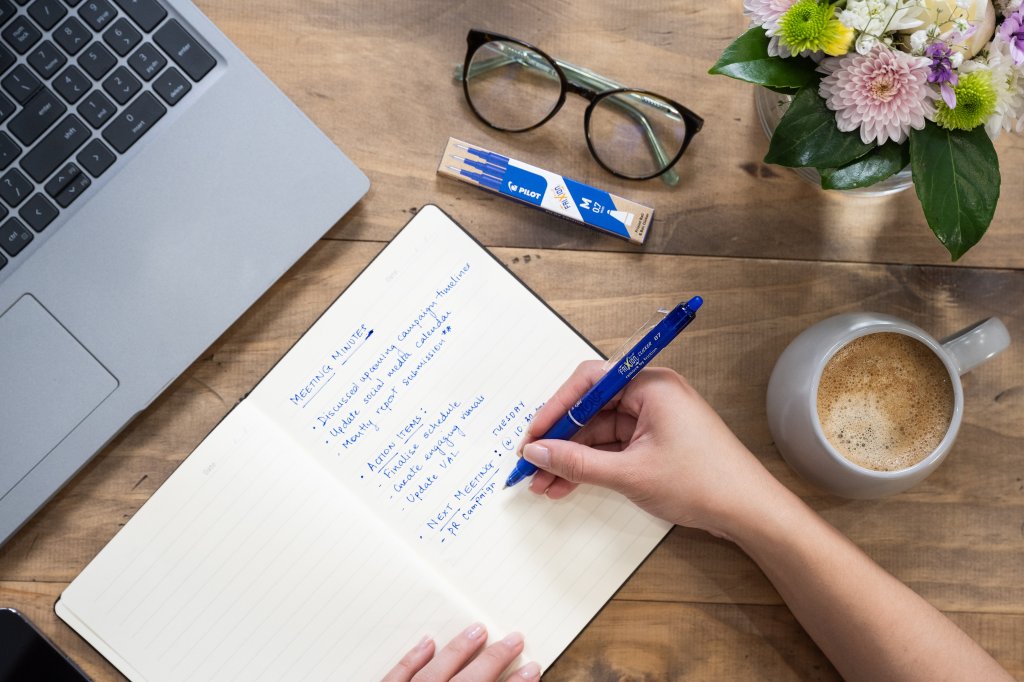 Person writing meeting notes in a notebook using a blue refillable pen, with a laptop, coffee, flowers, and a Pilot refill pack on the wooden desk.