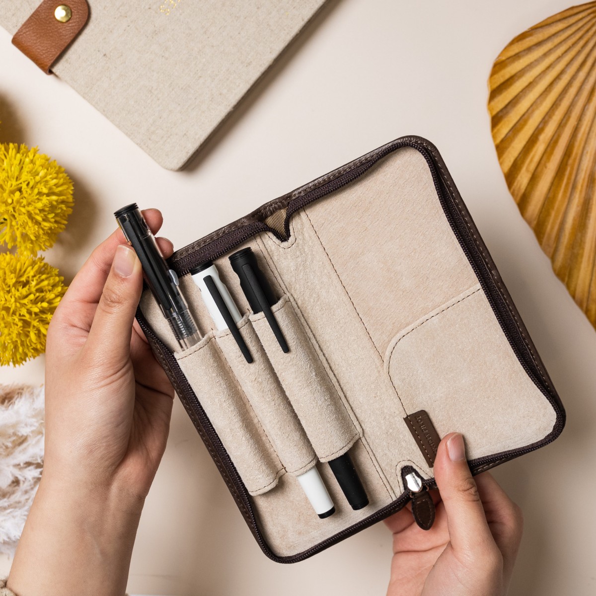 Three Pilot Explorer fountain pens in different colour barrels, a clear, white and black, sit in a Pilot Pensemble leather case, showcasing the collectable nature of fountain pens. A notebook with a linen cover can be seen in the top of the image. On the bottom right corner is a bottle of Pilot iroshizuku ink in deep pink, called kosumosu, which translates to cosmos in Japanese, a pink flower that looks similar to a daisy.