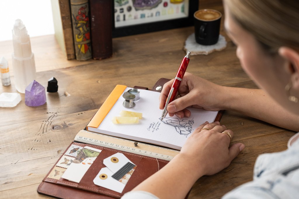 Woman writing with orange Capless SE fountain pen by PILOT Pen, in a notepad on a desk.