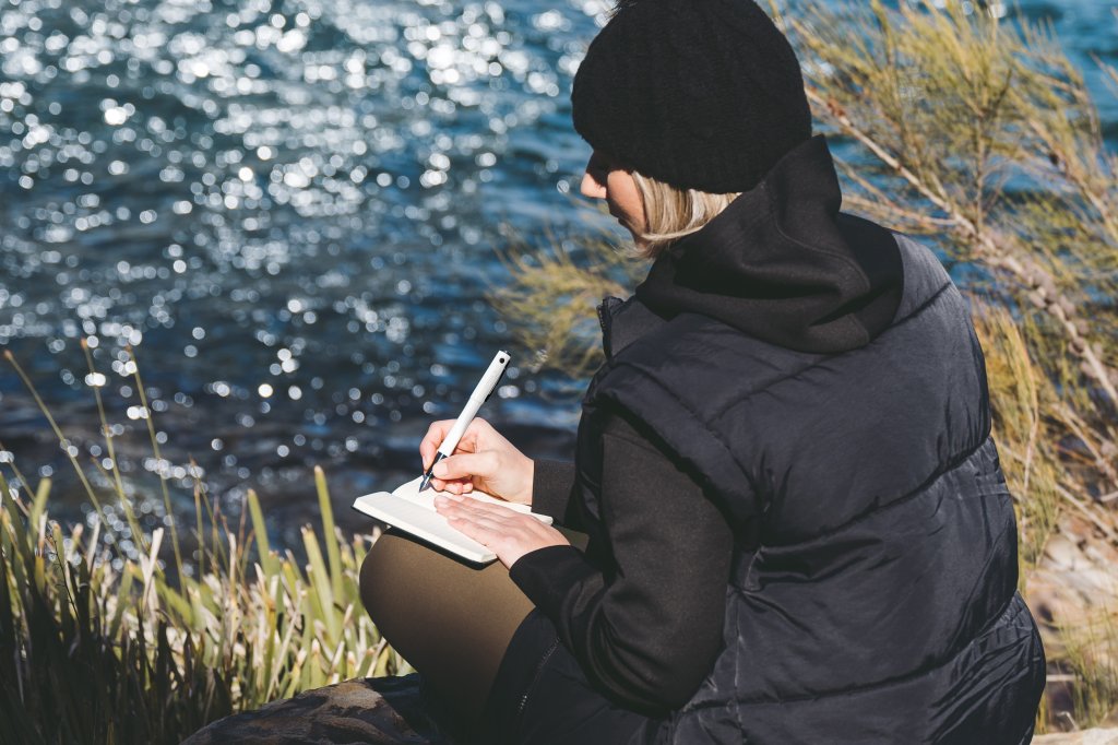 Woman sitting on a rock near a lake journaling with her fountain Pilot Pen.