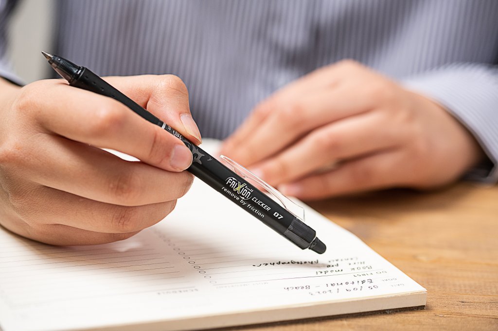 Close-up of a person writing in a planner with a black FriXion Clicker erasable pen, embracing pen and paper for daily productivity.