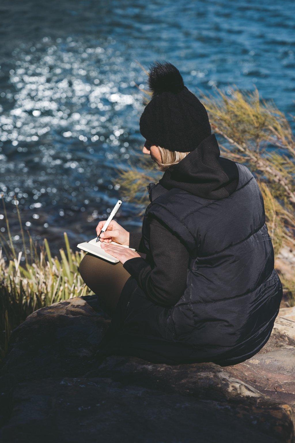 Woman journaling with a pen and paper by a lake, embracing an analogue break from screen time.