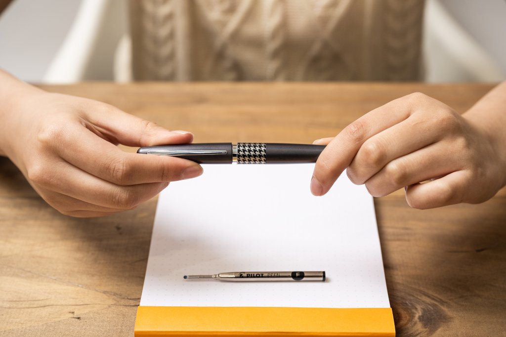 Person holding a refillable pen above a notepad with a Pilot pen refill placed on the paper.
