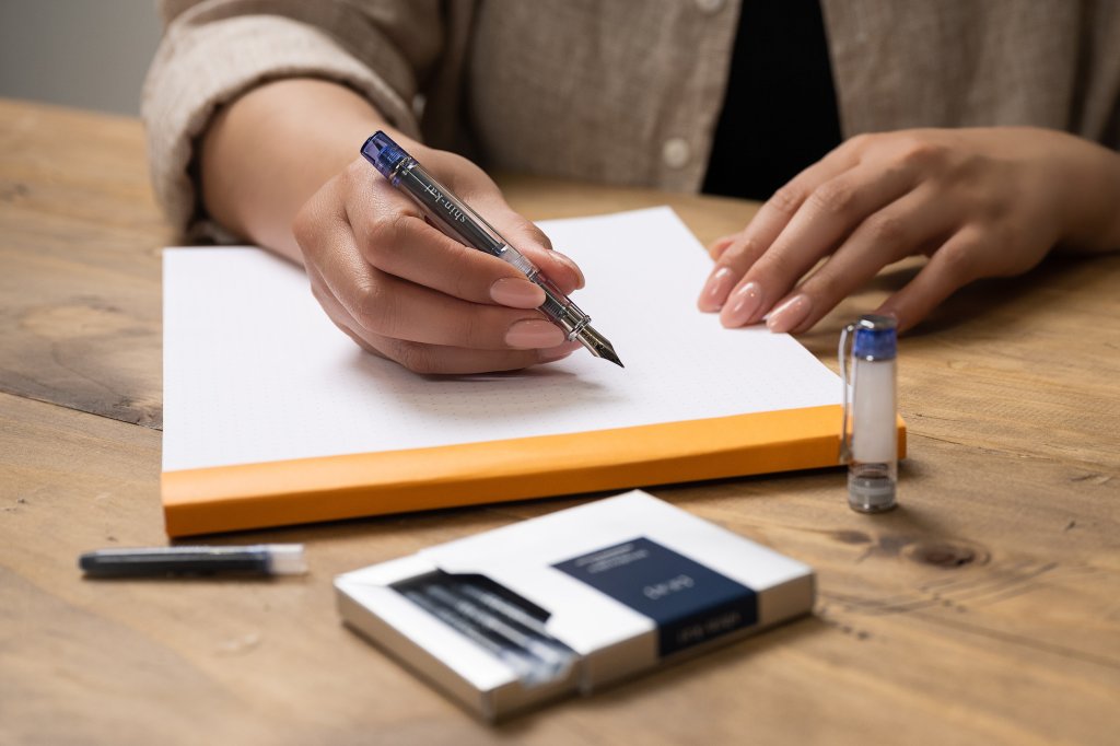 Pilot Prera Transparent pen fountain pen writing on notepad with blue black ink. The writer is sitting on a wooden table and in the foreground, there is an open pack of Pilot iroshizuku ink cartridges in blue black ink, called shin-kai, which translates to Deep Sea in Japanese.